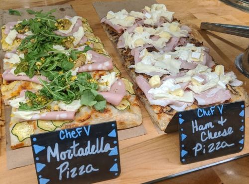 Two types of pizza on a wooden surface: Mortadella pizza with arugula and Ham & Cheese pizza, both labeled.
