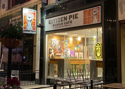 Exterior of Citizen Pie Roman Cafe at night, featuring a large window with artwork and a sign displaying the cafe's name.
