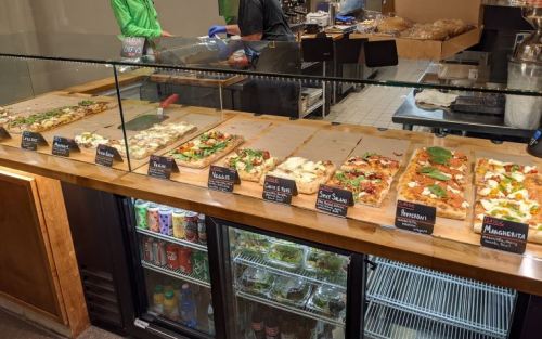 A display case with various types of pizza and salads, labeled with names, in a casual dining setting.