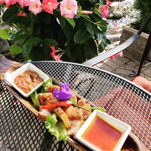 A wooden platter with sliced meat, vegetables, and two sauces, set on a table with pink flowers in the background.