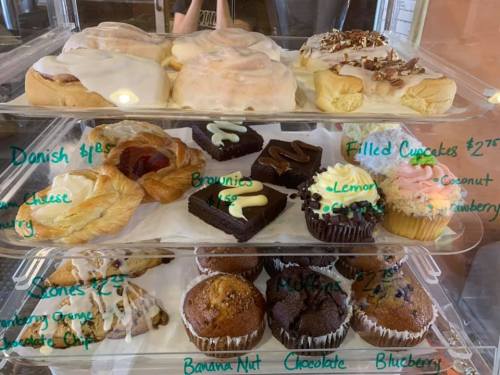 A display case filled with various baked goods, including danishes, brownies, cupcakes, and muffins, with prices listed.