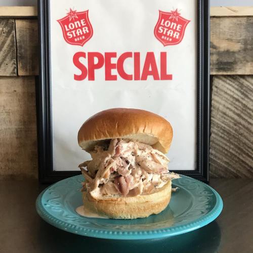 A chicken sandwich with shredded meat on a bun, placed on a blue plate in front of a "Lone Star Beer Special" sign.