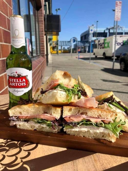 A sandwich stacked with greens and ham, served with a bottle of Stella Artois, against a sunny outdoor backdrop.
