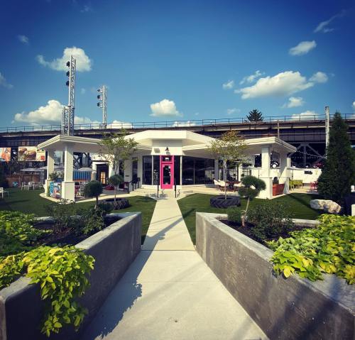 A modern building with a pink door, surrounded by greenery and planters, under a blue sky with clouds.