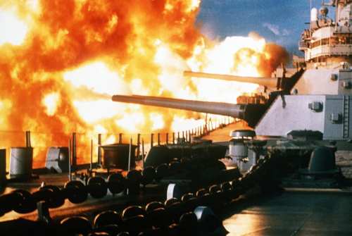 A battleship fires its cannons, creating a large explosion and billowing flames in the background.