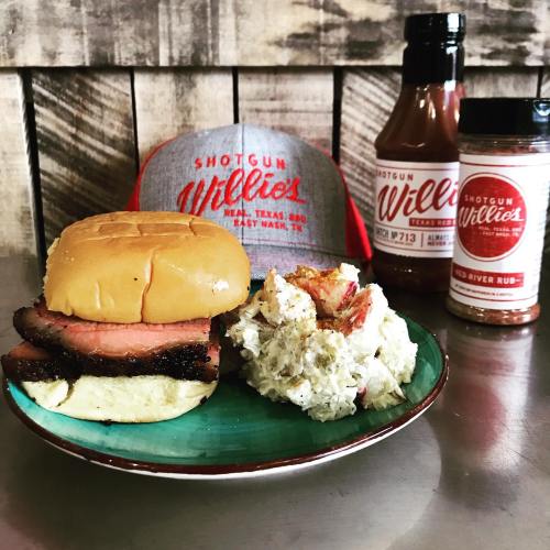 A plate with a brisket sandwich, potato salad, and Shotgun Willies hat and sauces in the background.
