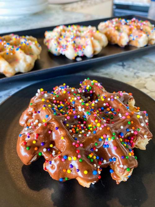 A chocolate-covered waffle topped with colorful sprinkles, served on a black plate, with more waffles in the background.