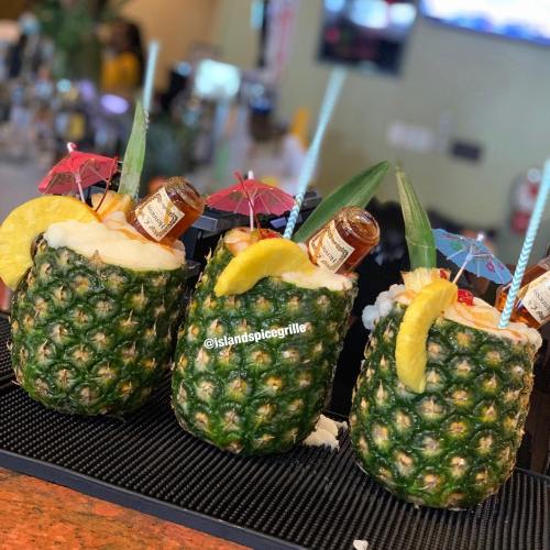 Three pineapples filled with tropical drinks, garnished with umbrellas, fruit, and bottles, sitting on a bar counter.