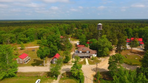Aerial view of a rural area with several buildings, red roofs, a water tower, and lush green trees surrounding the landscape.