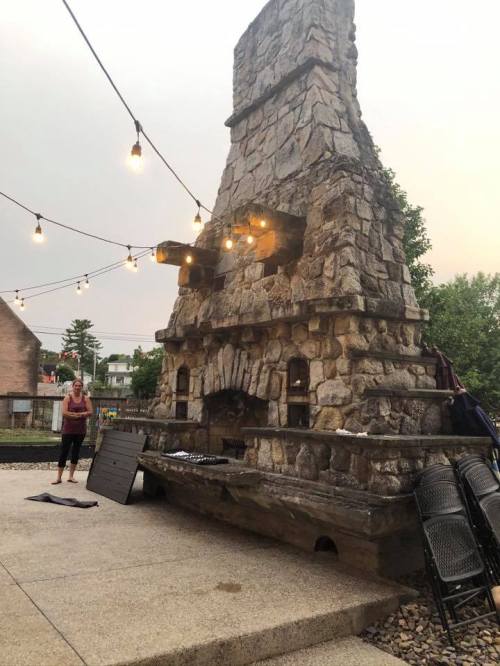 A large stone outdoor fireplace with string lights, surrounded by gravel and empty chairs, on a cloudy day.