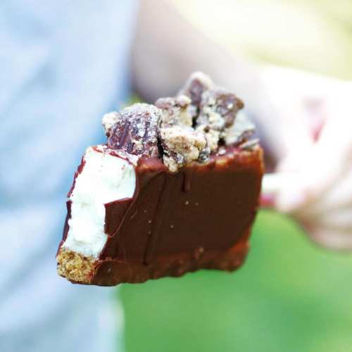 A close-up of a chocolate-covered ice cream bar with cookie pieces on top, held by a person outdoors.