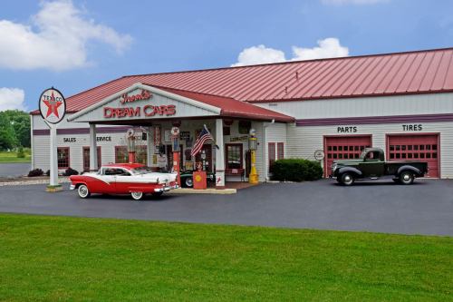 A vintage car dealership with a red classic car and an old truck outside, featuring a retro gas station sign.