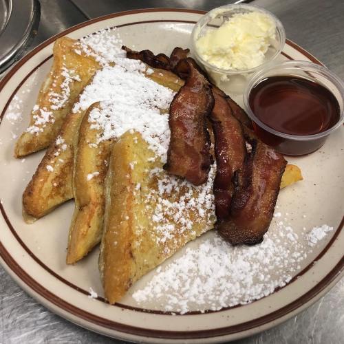 A plate of French toast topped with powdered sugar, crispy bacon, butter, and syrup on the side.