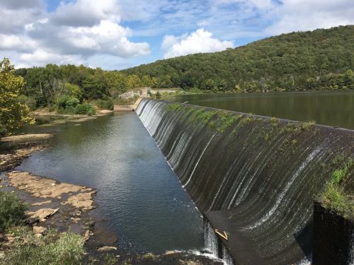 A serene landscape featuring a dam with water cascading over it, surrounded by lush greenery and a calm lake.