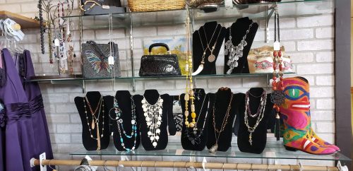 Display of various necklaces and accessories on black busts, with colorful bags and a decorative boot in the background.