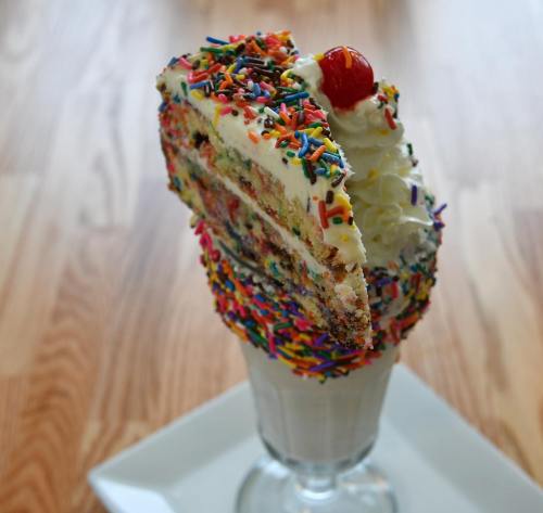 A tall glass filled with a milkshake topped with a slice of colorful cake, whipped cream, sprinkles, and a cherry.