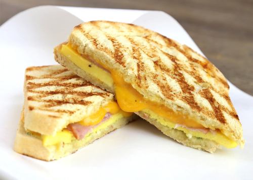 Grilled sandwich with melted cheese, ham, and egg, cut in half, served on a white plate.