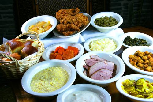 A spread of Southern comfort food, including fried chicken, greens, ham, and various sides in white bowls.