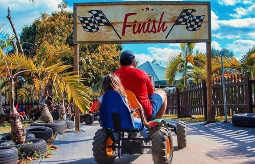 A person and a child ride a pedal tractor towards a "Finish" sign, surrounded by palm trees and tires.