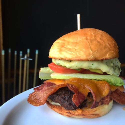 A close-up of a juicy burger with bacon, cheddar cheese, lettuce, tomato, and a creamy sauce on a toasted bun.