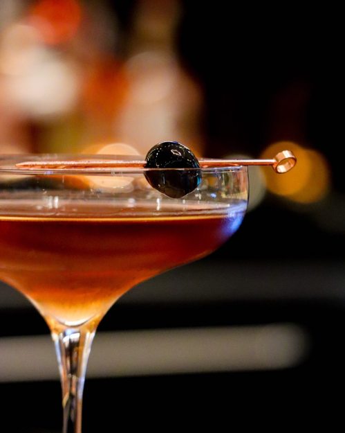 A close-up of a cocktail in a glass, garnished with an olive on a decorative skewer, with a blurred background.