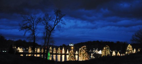 A serene night scene featuring illuminated Christmas trees reflected in a calm water body under a cloudy sky.