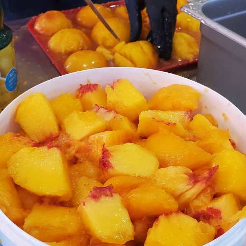 A bowl of freshly cut yellow peaches, with a person slicing more peaches in the background.