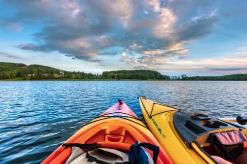 Two kayaks, one orange and one yellow, float on a calm lake under a colorful sky with clouds and green hills in the background.