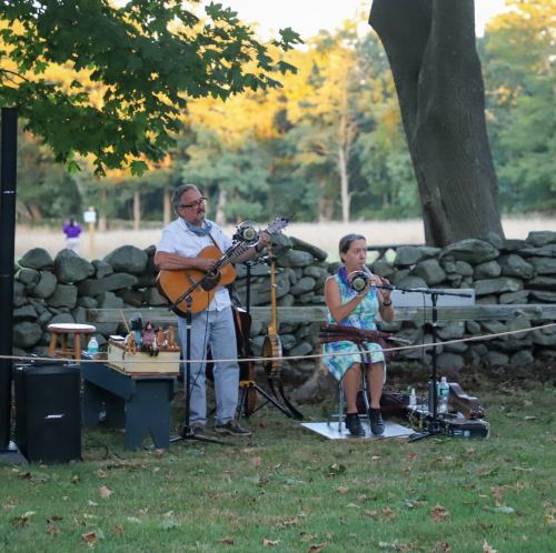 A man plays guitar while a woman plays flute, performing outdoors near a stone wall with trees in the background.