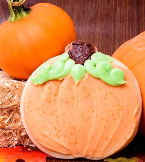 A large pumpkin-shaped cookie with orange frosting and green icing, surrounded by small pumpkins and hay.