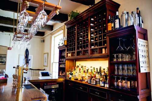 A wooden bar with shelves of wine bottles, glasses hanging above, and various spirits displayed on the counter.