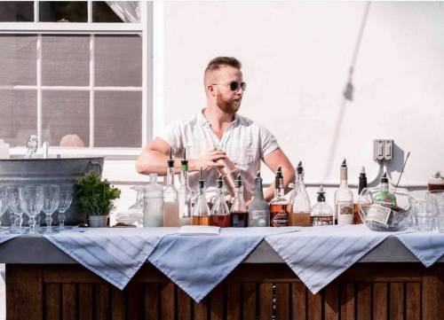 A bartender in sunglasses mixes drinks at a stylish outdoor bar with various bottles and glasses displayed.