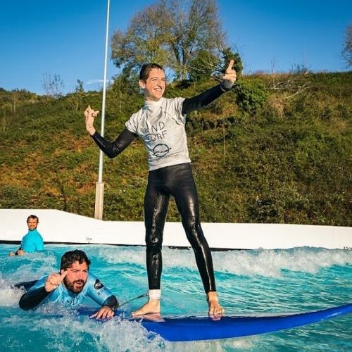A person balances on a surfboard while another swims nearby, both enjoying a sunny day at a surf park.