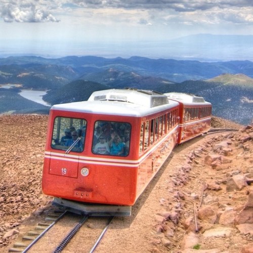 A red mountain train travels along a rocky track with scenic mountains and a lake in the background.