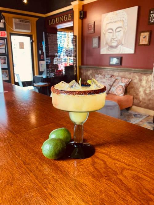 A vibrant margarita in a glass sits on a wooden bar, with two limes beside it and a cozy lounge in the background.