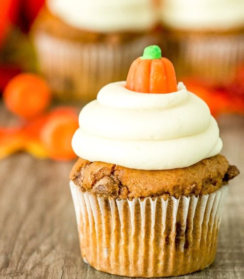 A close-up of a pumpkin cupcake topped with cream frosting and a small candy pumpkin decoration.