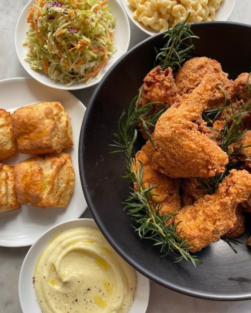 A bowl of crispy fried chicken garnished with rosemary, surrounded by sides of coleslaw, macaroni, and biscuits.