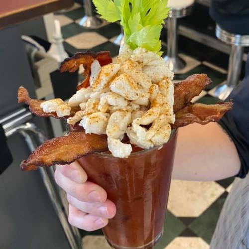 A hand holding a tall glass filled with a cocktail topped with bacon, chicken, and a celery stick.