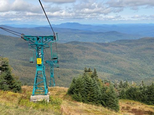 A ski lift tower stands against a backdrop of rolling mountains and a cloudy sky, surrounded by autumn foliage.