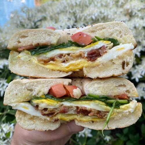 A hand holding a sandwich filled with eggs, bacon, spinach, and tomatoes, set against a backdrop of white flowers.