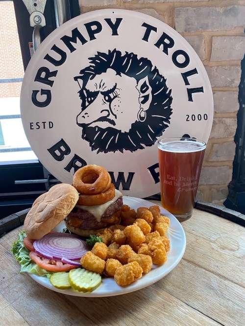 A hearty burger topped with onion rings, served with tater tots, pickles, and a pint of beer, against a brewery backdrop.