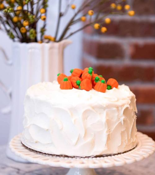 A white frosted cake topped with small orange and green pumpkin decorations, displayed on a cake stand.