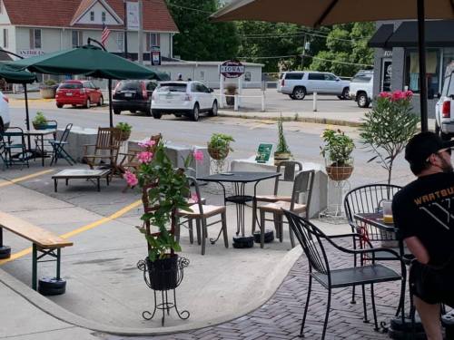 Outdoor café scene with tables, chairs, and potted flowers; cars visible in the background.