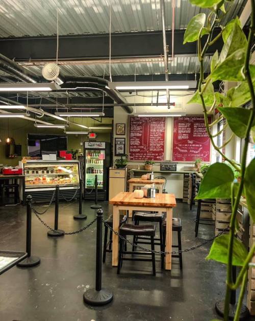 A cozy café interior with a wooden table, plants, and a menu board on the wall, featuring a display case of food.