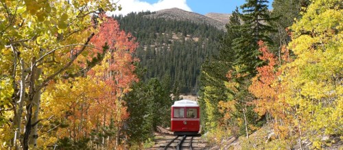 A red train travels along a scenic track surrounded by vibrant autumn foliage and mountains in the background.