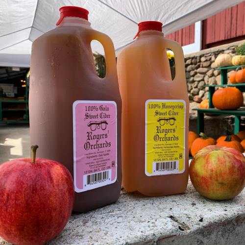 Two jugs of sweet cider labeled "Gala" and "Honeycrisp" next to a red apple, with pumpkins in the background.
