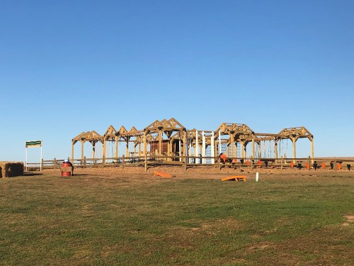 A wooden structure on a grassy field under a clear blue sky, with hay bales and seating areas nearby.