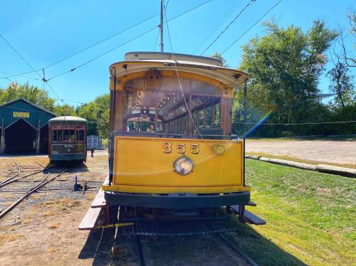 A vintage yellow streetcar numbered 355, parked on tracks with greenery in the background under a clear blue sky.