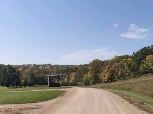A dirt road leads through a scenic landscape with trees displaying autumn colors under a clear blue sky.