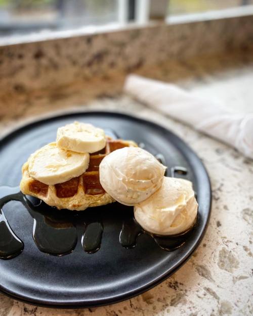 A black plate with a waffle topped with whipped cream and two scoops of ice cream, drizzled with syrup.
